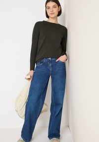 Dark green long-sleeve top paired with wide-leg blue jeans. The jeans feature a front pocket and subtle fading. A light beige bag is draped over her arm.
