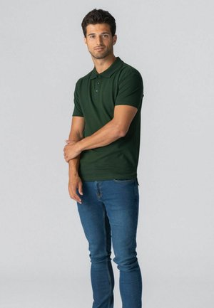 Mand stående med armene krydset, iført en mørkegrøn polotrøje og blå jeans mod en ensfarvet lys baggrund.