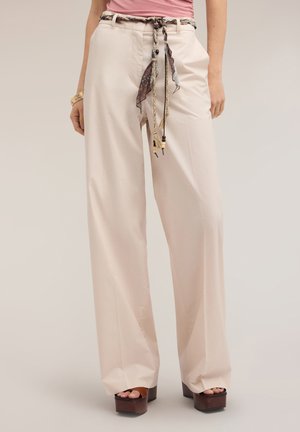 Vrouw draagt beige wijde broek met een patroon stoffen riem, bruine plateauzandalen en een roze top, staand tegen een effen achtergrond.