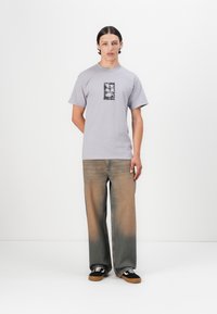 Lys grå bomull T-skjorte med et svart grafisk trykk i midten, kombinert med løspassede denimjeans med en gradient brun nyanse, og svarte sneakers.