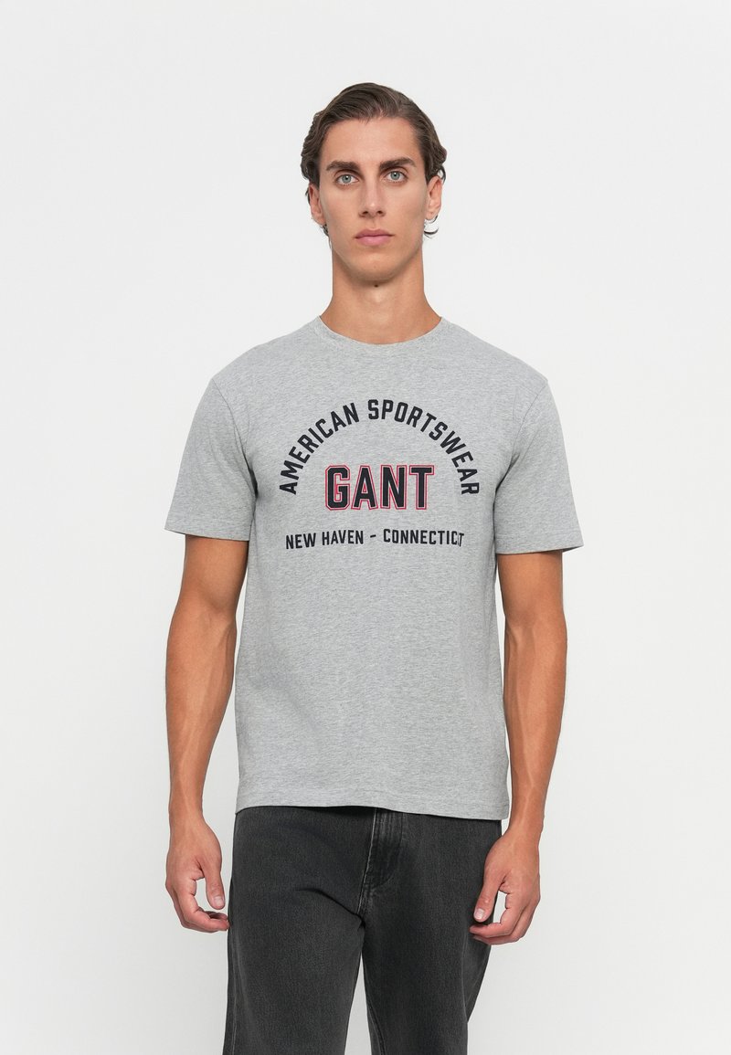 Ung mann iført en grå GANT T-skjorte med teksten "American Sportswear New Haven Connecticut", kombinert med svarte bukser, står mot en hvit bakgrunn.
