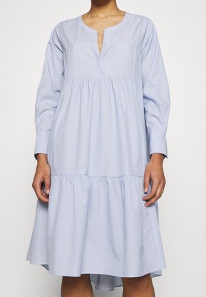 Woman wearing a light blue, long-sleeve, tiered dress with a V-neck slit and loose fit, standing against a plain background.