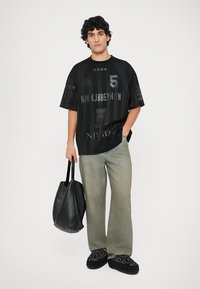 Løs passform svart grafisk t-skjorte med logoer og tall, kombinert med lysegrå jeans, svart veske og broderte svarte tøfler.