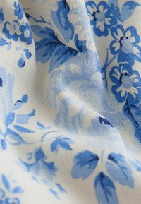 Lengvas audinys su mėlynais gėlių ir lapų raštais, matyti raukšlės ir austos medžiagos tekstūra.