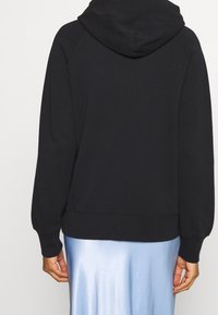 Felpa nera con cappuccio, con polsini e orlo a coste, maniche lunghe e vestibilità rilassata. Indossata sopra una gonna blu lucida. Tessuto semplice e testurizzato.