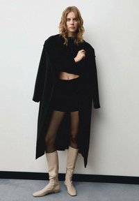 Černý dlouhý kabát, černý crop top a kraťasy s průhlednými punčochami; krémové vysoké boty. Hladké materiály, elegantní design a kontrastní barvy.