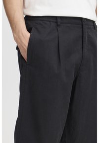 Black trousers made of soft fabric, featuring a pleated design, side pockets, a buttoned waistband, and a loose fit.
