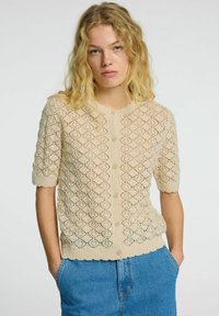 Beige pletený kardigan s krátkým rukávem, ozdobený diamantovým vzorem, volány na spodní části a zapínáním na knoflík. Nosený přes modré džíny.