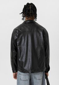 Zwarte leren jas met een gladde textuur, gestructureerd ontwerp en lange mouwen. Voorzien van een niet-vals handschoen en een rechte zoom.