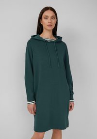 Vrouw in een donkergroene met capuchon jurk over een gestreepte lange mouwen shirt, staand tegen een effen grijze achtergrond.