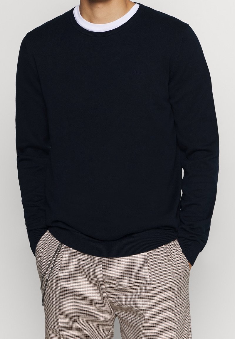 Marineblauer Strickpullover mit rundem Halsausschnitt, gerippten Bündchen und Saum. Kombiniert mit hellen karierten Hosen.