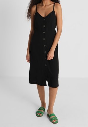 Frau trägt ein schwarzes, knöpfiges Midikleid mit Spaghettiträgern und grünen offenen Sandalen und steht vor einem schlichten Hintergrund.