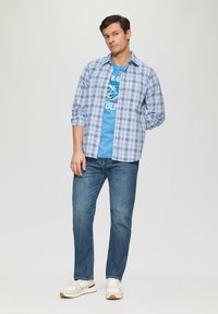 Blauwe en witte geruite langemouwen shirt over een blauwe graphic t-shirt, in combinatie met blauwe jeans en lichte sneakers.