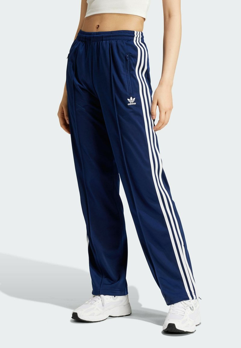 Pantalones de chándal azul marino fabricados con material ligero, que presentan franjas laterales blancas, una cinturilla elástica y bolsillos con cremallera.