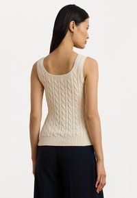 Top de malha em ponto corrido bege com costas em forma de cavalo, bainha canelada e alças largas. A textura apresenta padrões torcidos em relevo por toda a peça.