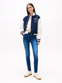 Marineblå og hvit varsityjakke med logo, kremfarget genser, blå skinnyjeans og hvite sneakers med gummisåle. En enkel, uformell antrekk.