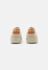 Sneaker bianche con accenti in suede beige e linguette arancioni sul tallone. Le calzature presentano il marchio "VEJA" stampato in bianco sul tallone e una suola testurizzata.
