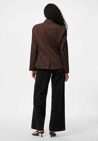 Blazer marron ajusté avec une fente unique, associé à un jean noir à jambes larges et des sandales à talons. Tissu lisse et silhouette structurée.