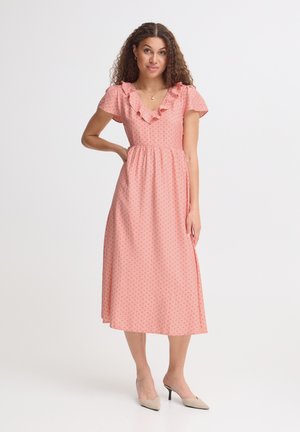 Femme aux cheveux bouclés portant une robe midi rose avec col volanté et petit motif à pois, debout avec une main sur la hanche et des talons beiges.