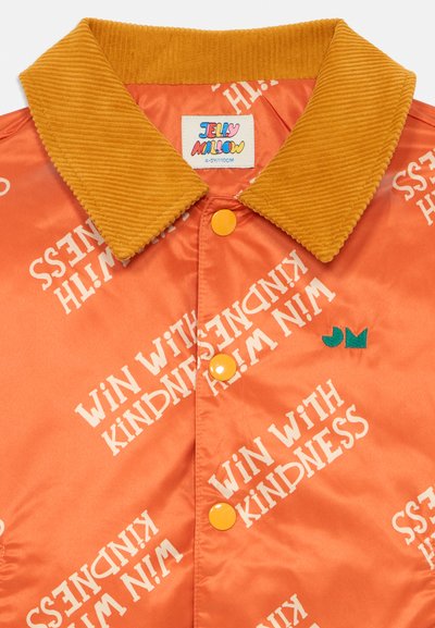 Orange Samtjacke mit gelbem Cordkragen, mit "WIN WITH KINDNESS" in Weiß bedruckt, gelben Knöpfen und einem grünen Logo.
