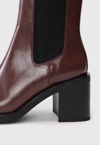 Bottine en cuir marron avec panneau latéral élastique noir et talon bloc noir épais sur fond clair.