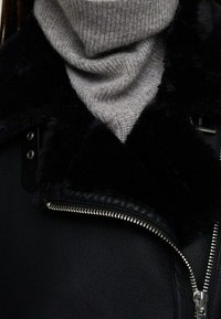 Veste en cuir noir avec un col en fausse fourrure, dotée d'une fermeture éclair en argent et d'une écharpe grise côtelée, offrant un contraste de texture et de chaleur.