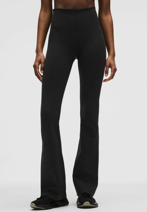 Femme portant un pantalon évasé noir taille haute et des baskets noires, debout devant un fond gris clair uni.