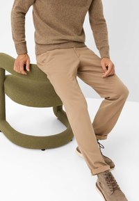 Beige chinos met een rechte snit, gecombineerd met een bruine trui en bruine veterschoenen, zittend op een groen gestoffeerde stoel.