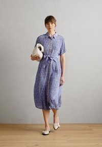 Vestido azul com padrão, mangas curtas e cintura com laço, apresentando uma parte frontal com botões e um comprimento abaixo do joelho. Modelo segura uma mala creme.