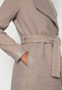 Beige ullblandad kappa med bred krage, bälte i midjan och läderdetaljer vid ärmsluten. Mjuk textur med en subtil fiskbensmönster.