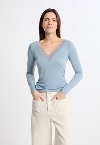 Light blue long-sleeve top with a V-neck, featuring a delicate lace trim along the neckline. Soft, fitted fabric complements wide white pants.