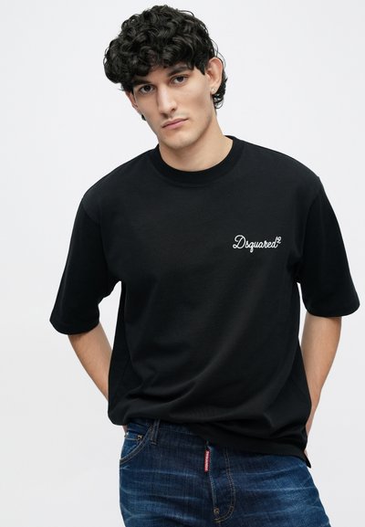 Melnā kokvilnas T-krekls ar īsām piedurknēm un nelielu balto "Dsquared²" logotipu krūtīs, kombinēts ar zilām džinsa biksēm.