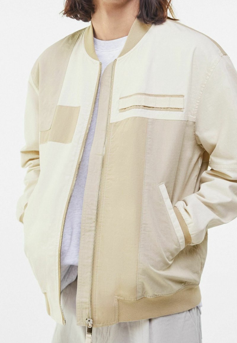 Jeune personne portant une veste zippée bicolore beige et crème avec poches mains et col et poignets côtelés.