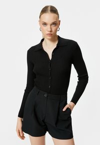 Schwarzes geripptes Langarmshirt mit einem geknöpften Kragen, kombiniert mit hochtaillierten schwarzen Falten-Shorts, die einen vorderen Knopfverschluss haben.