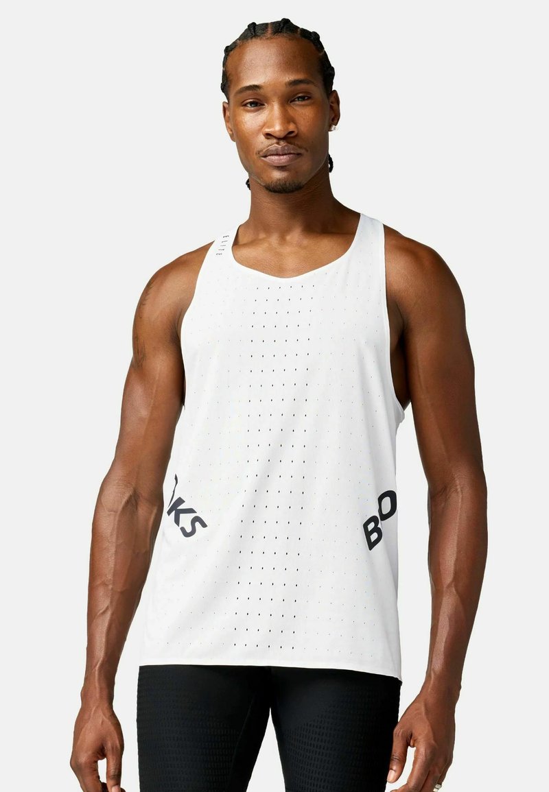 Hombre con el pelo trenzado, vistiendo una camiseta deportiva blanca sin mangas perforada y pantalones cortos negros ajustados, de pie frente a un fondo liso.
