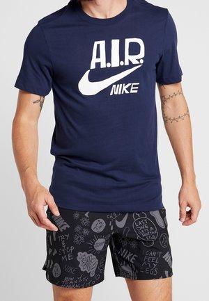 Mężczyzna noszący granatową koszulkę Nike A.I.R. i czarne spodenki Nike z szarymi nadrukami i rysunkami.