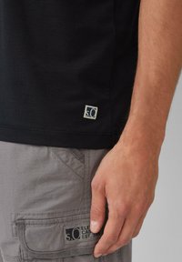 Czarna koszulka z krótkim rękawem i małą metką logo na dolnym brzegu. Połączona z szarymi spodniami cargo z kieszenią z naszywką marki.