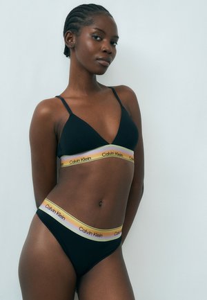 Femme portant un bralette noir Calvin Klein et une culotte assortie avec des bandes élastiques rayées arc-en-ciel, debout devant un fond clair uni.