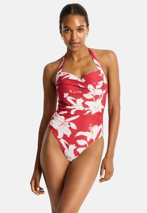 Kobieta w czerwonym kostiumie kąpielowym na szyję z dużym białym kwiatowym wzorem, stojąca na jednolitym jasnym tle.