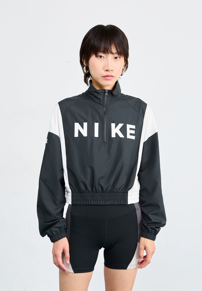 Veste noire et blanche avec un col zippé, présentant un grand logo "NIKE" à l'avant et une taille élastique, assortie avec un short noir.