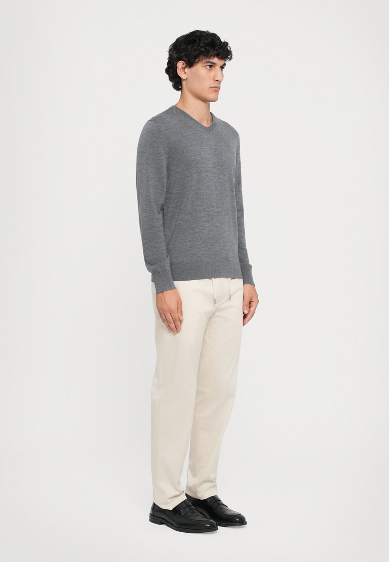 Grijze v-hals trui gemaakt van gebreide stof, gecombineerd met lichtbeige broeken. Het model draagt zwarte schoenen en staat tegen een effen witte achtergrond.