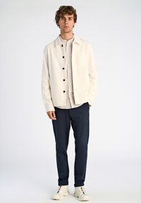 Ljust beige knappad jacka med svarta knappar över en randig skjorta. Marinblå byxor och vita sneakers fulländar outfiten.