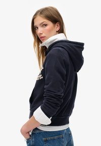 Marineblå hættetrøje med kængurulomme, broderet logo og snore. Laget over en hvid skjorte med krave og parret med blå jeans.