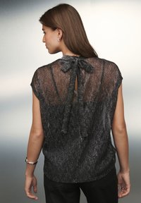 Mustast läbipaistvast metallilõngadega pluus lühikeste varrukatega ja tagant lipsuga. Silmatorkav on võtmeauguga avaus ja tekstuuriga disain.