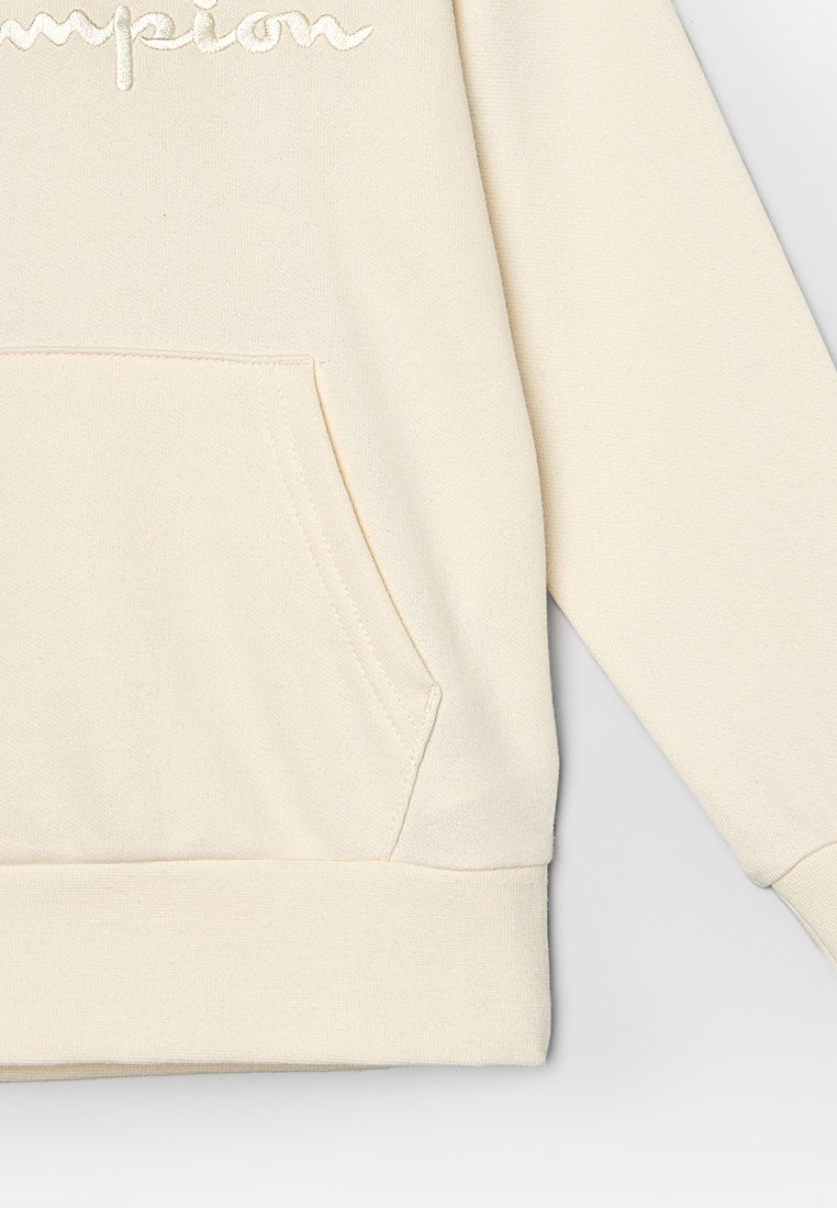 Sudadera color crema, material de algodón suave, presenta un bolsillo frontal y puños acanalados. Logo bordado de "Champion" en la parte superior del pecho.