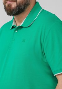 Polo vert en tissu texturé, avec un col classique doté d'une bordure blanche et d'un logo sur le côté gauche. Trois boutons au niveau du cou.