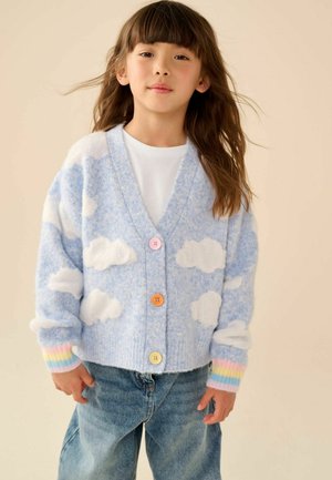 Young child with long brown hair wearing a blue cardigan with white clouds and multicolor striped cuffs, white shirt, and blue jeans.