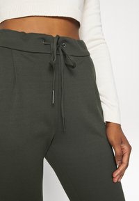 Tummanvihreät jogger-housut, jotka on valmistettu pehmeästä kankaasta. Niissä on joustava vyötärönauha, jonka nauhoissa on metalliset päätypidikkeet. Lähikuva näyttää käden asennon.