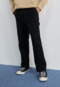 Zwarte broek van duurzaam materiaal, met een relaxte pasvorm en zijzakken, gecombineerd met zwart-witte sneakers op een blauwe vloer.