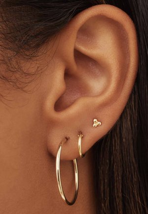 Boucles d'oreilles en or en forme de créole et une petite boucle d'oreille à trois perles. Gros plan sur une oreille, montrant une texture de peau lisse et des mèches de cheveux.